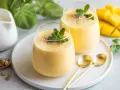 Dwa szklanki gęstej, kremowej pulpy mango, ozdobione listkami mięty i posypką. Obok świeży, pokrojony owoc mango.