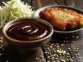 Miseczka z gęstym, ciemnym sosem tonkatsu, obok smażony kotlet i poszatkowana kapusta.