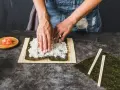 Ręce rozprowadzają ryż na nori, przygotowując zaprawę do sushi. Obok leżą pałeczki i marynowany imbir.