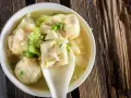 Aromatyczne pierożki wonton w bulionie, ozdobione szczypiorkiem i kawałkami warzyw, na drewnianym tle.