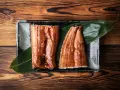 Dwa kawałki błyszczącego, glazurowanego unagi na liściu, podane na prostokątnej tacy na drewnianym tle.