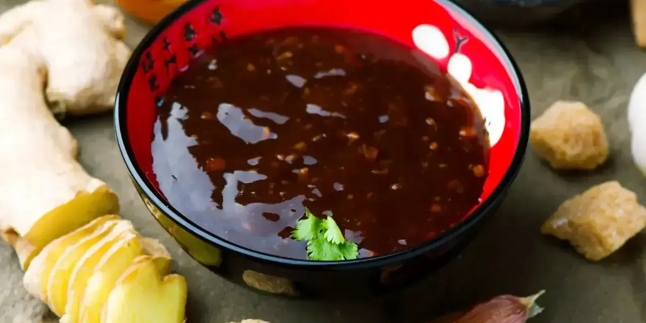 Miseczka z ciemnym sosem teriyaki, obok imbir, czosnek, cukier trzcinowy i miód.