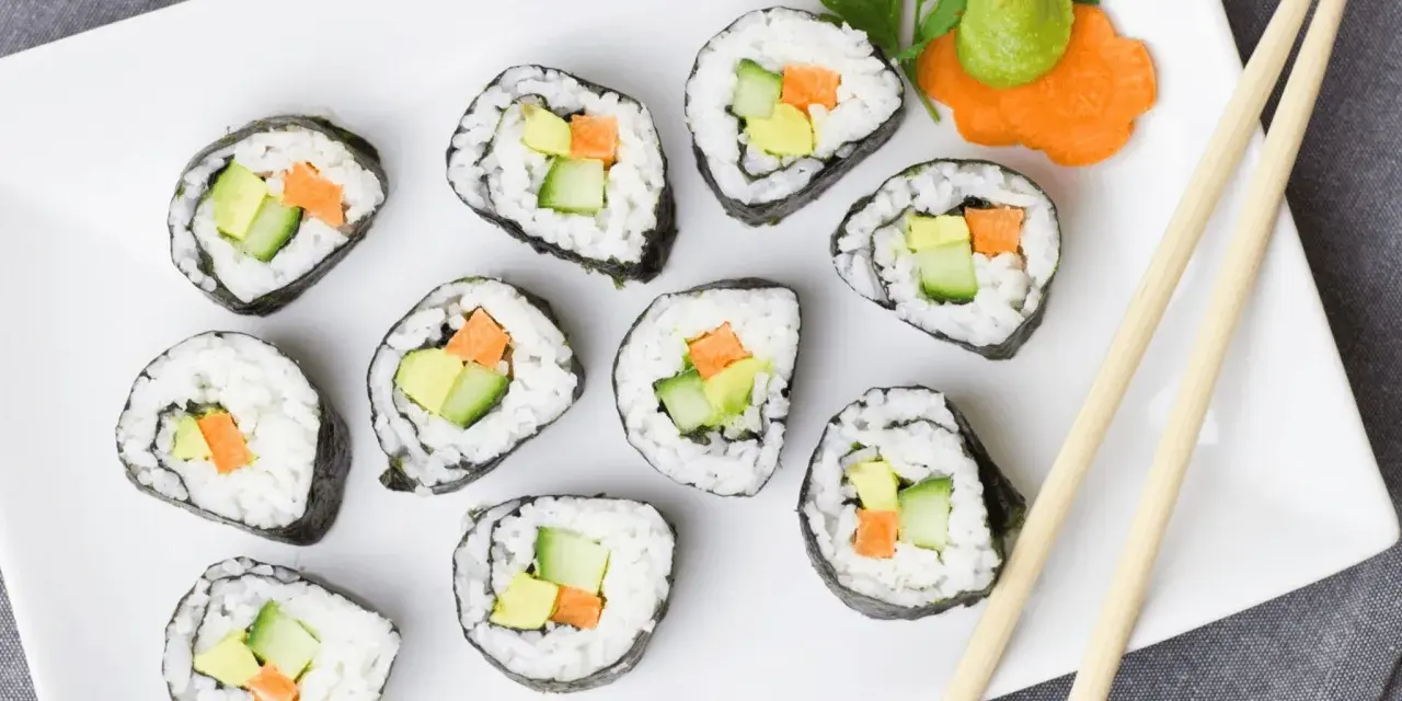 Talerz z rolkami sushi z awokado, ogórkiem i marchewką, pałeczki, wasabi i ozdobą z marchewki.