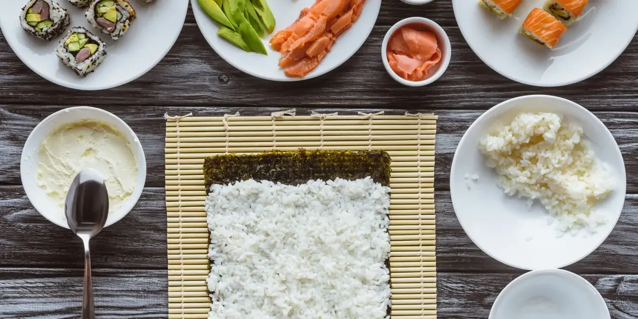 Przygotowanie sushi: ryż, nori, łosoś, awokado, wasabi i marynowany imbir na drewnianym blacie.