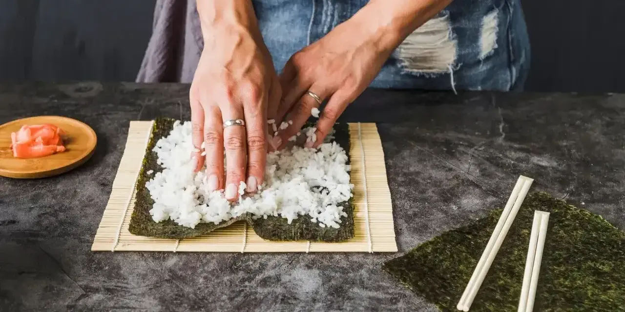 Ręce rozprowadzają ryż na nori, przygotowując zaprawę do sushi. Obok leżą pałeczki i marynowany imbir.