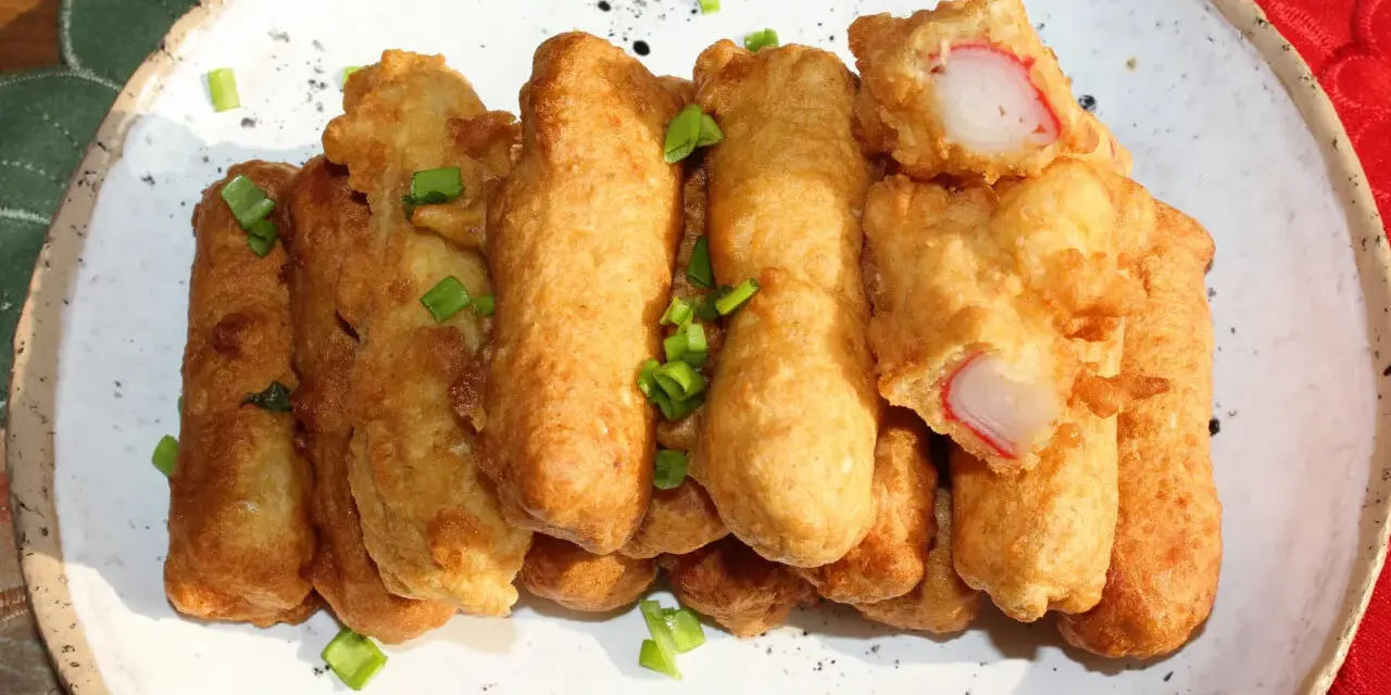 Chrupiące paluszki surimi w cieście, posypane szczypiorkiem, prezentują się apetycznie na białym talerzu.