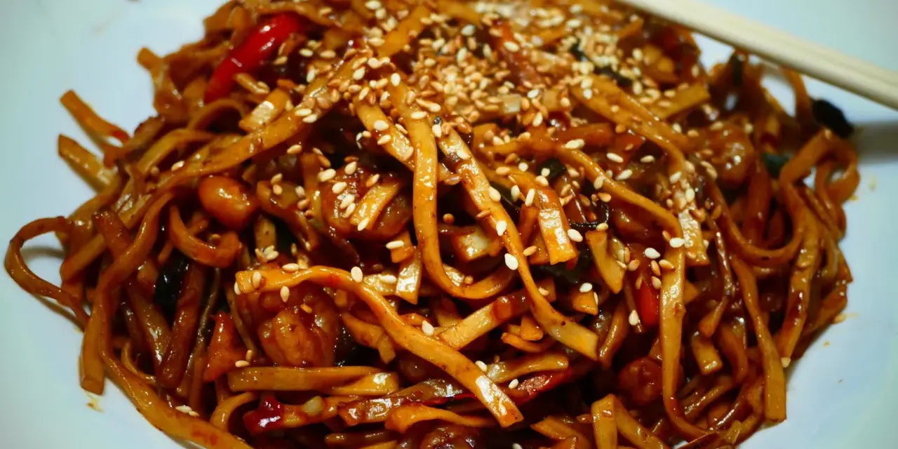 Makaron chow mein z sezamem i papryką na białym talerzu, z pałeczkami.