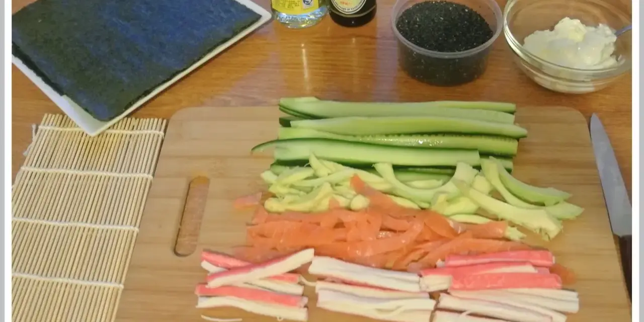 Pokrojone składniki do sushi: ogórek, łosoś, paluszki krabowe, awokado, sezam, sos sojowy, ryż i nori.