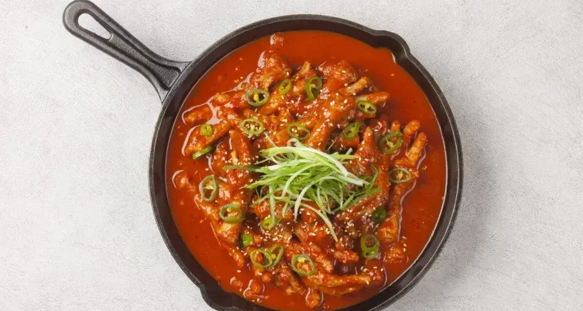 Kurczak w sosie gochujang, posypany sezamem i zieloną cebulką, podany w żeliwnej patelni.
