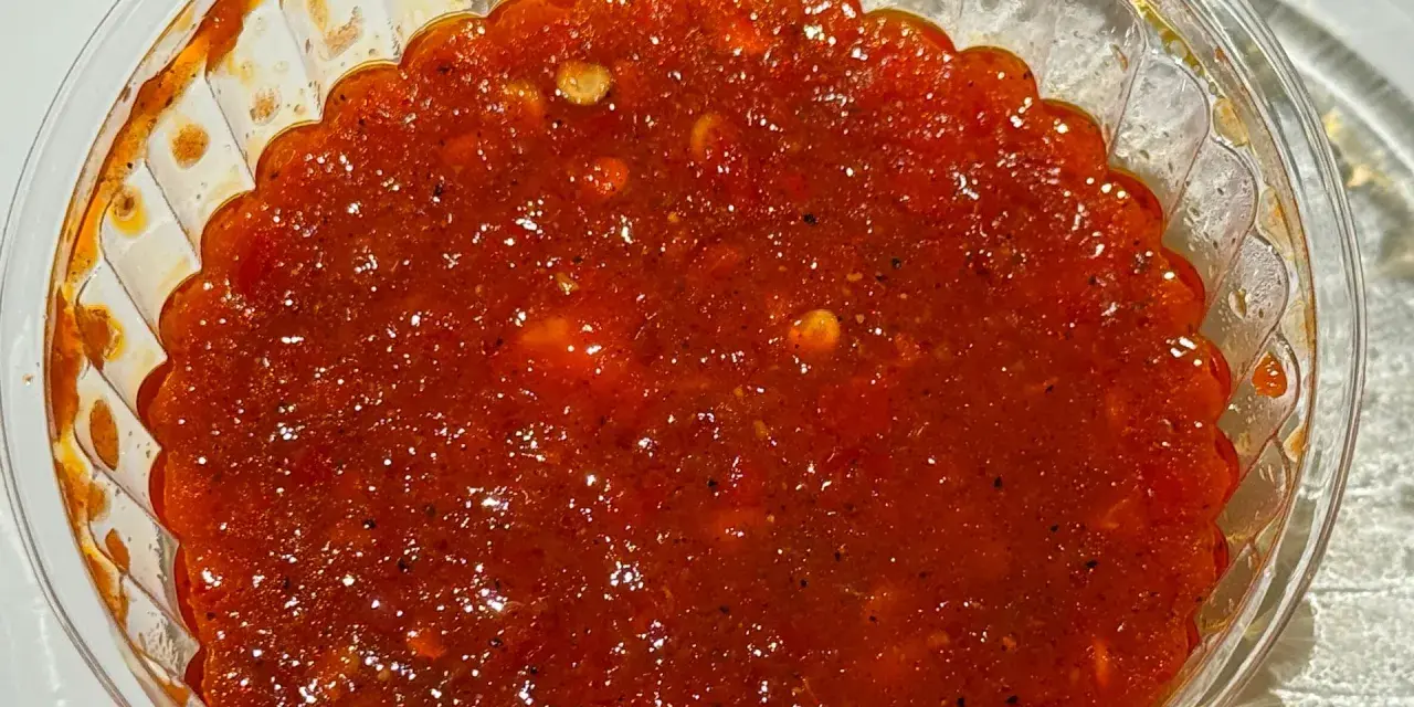 Gęsty, czerwony sos sambal w przezroczystym pojemniku, z widocznymi kawałkami papryczek chili i przypraw.