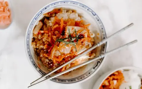 Miseczka ryżu z pikantnym kimchi i startą marchewką, ozdobiona listkami mięty. Obok pałeczki i mała miseczka z dodatkami, jak do koreańskiego banchan.