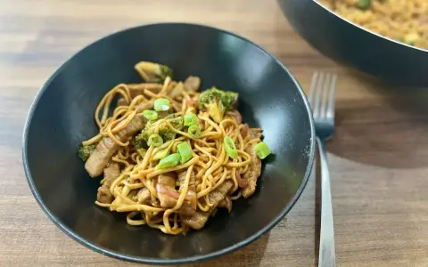 Pyszny stir fry z wołowiną, makaronem i brokułami, posypany szczypiorkiem. Obok widać patelnię z daniem.