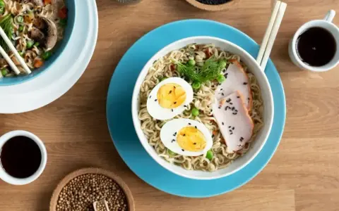 Miseczka tantanmen z makaronem, jajkiem, plastrami mięsa i zielonym koperkiem. Obok sos sojowy i ziarna.