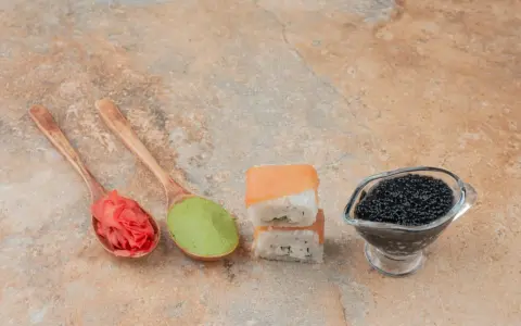 Na drewnianych łyżkach różowy imbir do sushi i zielone wasabi. Obok dwa kawałki sushi i sos sojowy z kawiorem.
