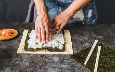 Ręce rozprowadzają ryż na nori, przygotowując zaprawę do sushi. Obok leżą pałeczki i marynowany imbir.