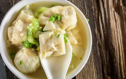 Aromatyczne pierożki wonton w bulionie, ozdobione szczypiorkiem i kawałkami warzyw, na drewnianym tle.