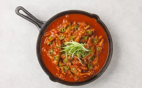 Kurczak w sosie gochujang, posypany sezamem i zieloną cebulką, podany w żeliwnej patelni.