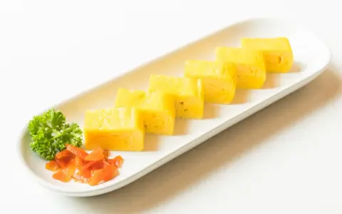 Sześć kawałków puszystego omletu tamago ułożonych na białej tacy, ozdobionej natką pietruszki i pomarańczowym dodatkiem.