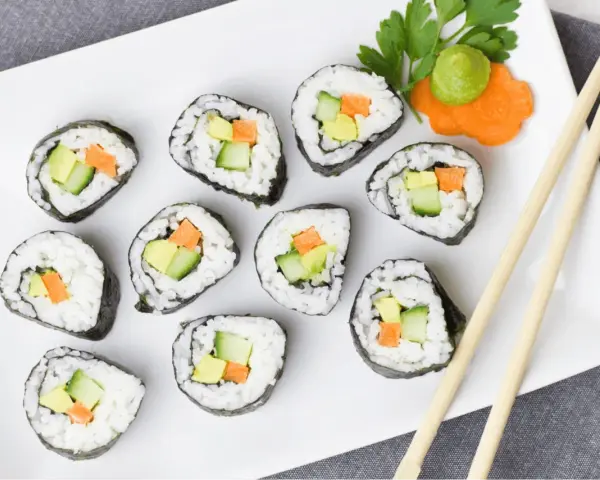 Talerz z rolkami sushi z awokado, ogórkiem i marchewką, pałeczki, wasabi i ozdobą z marchewki.