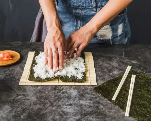 Ręce rozprowadzają ryż na nori, przygotowując zaprawę do sushi. Obok leżą pałeczki i marynowany imbir.