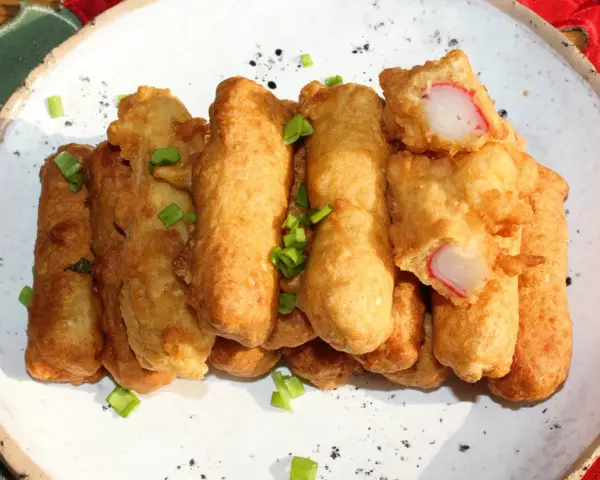 Chrupiące paluszki surimi w cieście, posypane szczypiorkiem, prezentują się apetycznie na białym talerzu.