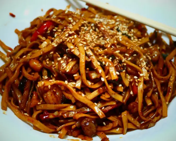 Makaron chow mein z sezamem i papryką na białym talerzu, z pałeczkami.