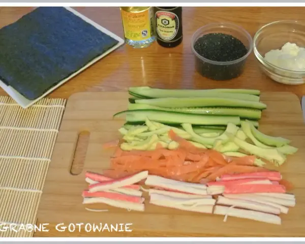 Pokrojone składniki do sushi: ogórek, łosoś, paluszki krabowe, awokado, sezam, sos sojowy, ryż i nori.
