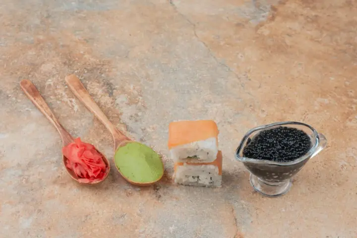Na drewnianych łyżkach różowy imbir do sushi i zielone wasabi. Obok dwa kawałki sushi i sos sojowy z kawiorem.