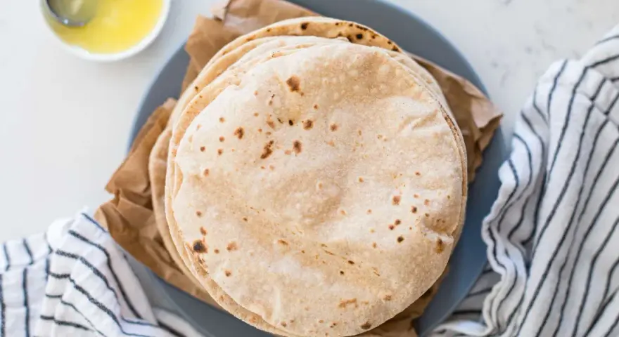 Stos świeżo upieczonych, lekko przypieczonych roti na talerzu, obok miseczki z roztopionym masłem.