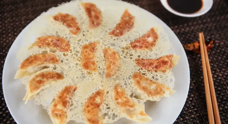 Chrupiące pierożki gyoza ułożone na talerzu, z chrupiącą, złotą panierką. Obok miseczka sosu sojowego i pałeczki.