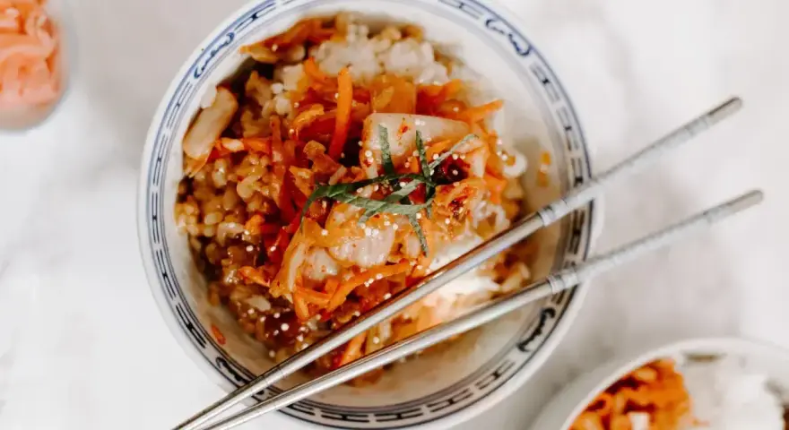 Miseczka ryżu z pikantnym kimchi i startą marchewką, ozdobiona listkami mięty. Obok pałeczki i mała miseczka z dodatkami, jak do koreańskiego banchan.
