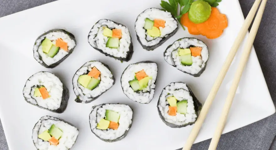 Talerz z rolkami sushi z awokado, ogórkiem i marchewką, pałeczki, wasabi i ozdobą z marchewki.