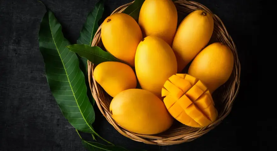 Koszyk pełen soczystych, żółtych mango, jedno przekrojone w kostkę. Obok zielone liście mango.