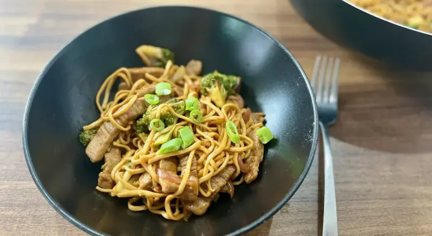 Pyszny stir fry z wołowiną, makaronem i brokułami, posypany szczypiorkiem. Obok widać patelnię z daniem.