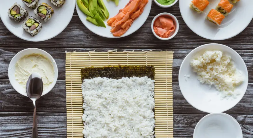 Przygotowanie sushi: ryż, nori, łosoś, awokado, wasabi i marynowany imbir na drewnianym blacie.