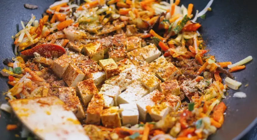 Kawałki tofu i kolorowe warzywa po chińsku smażą się na patelni wok, przyprawione kurkumą i papryką.