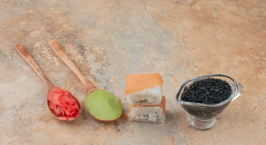Na drewnianych łyżkach różowy imbir do sushi i zielone wasabi. Obok dwa kawałki sushi i sos sojowy z kawiorem.