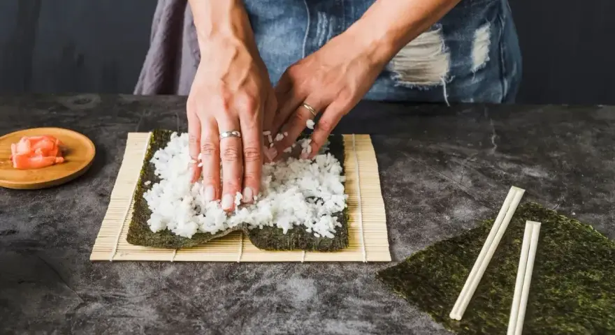 Ręce rozprowadzają ryż na nori, przygotowując zaprawę do sushi. Obok leżą pałeczki i marynowany imbir.