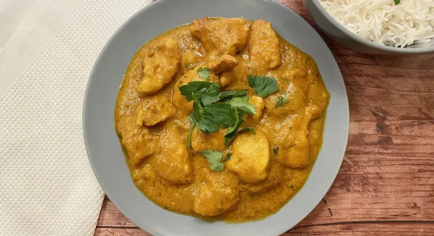 Aksamitny kurczak po indyjsku w kremowym sosie, udekorowany świeżą kolendrą, obok miseczki ryżu basmati.