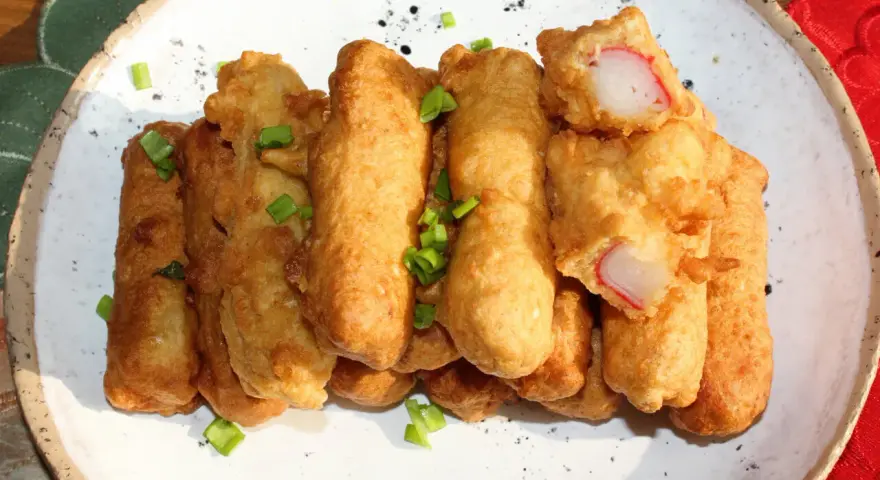Chrupiące paluszki surimi w cieście, posypane szczypiorkiem, prezentują się apetycznie na białym talerzu.