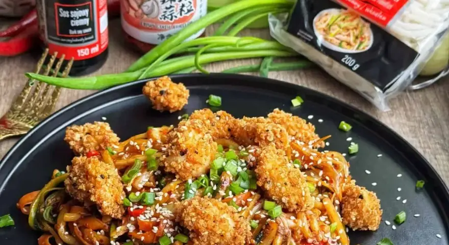 Pyszny makaron udon z kurczakiem w panierce i kolorowymi warzywami, posypany sezamem i szczypiorkiem.