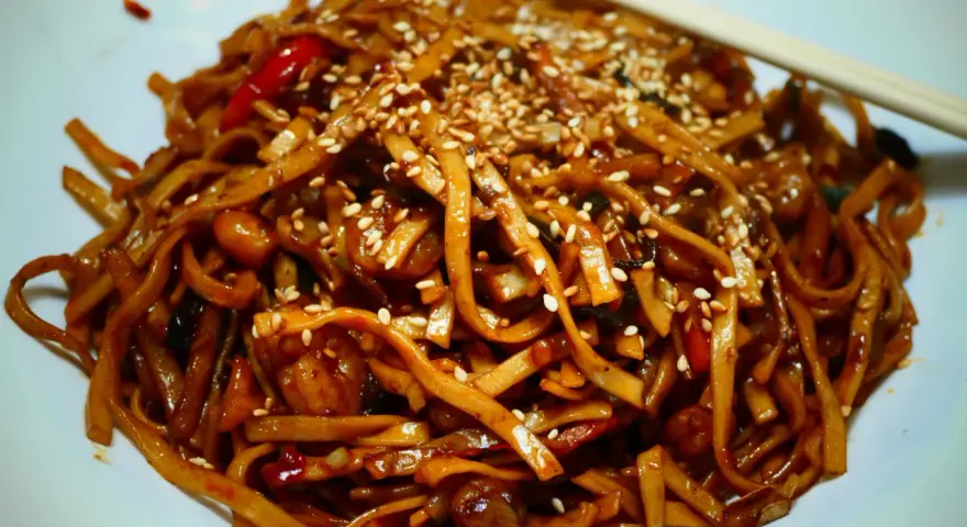 Makaron chow mein z sezamem i papryką na białym talerzu, z pałeczkami.