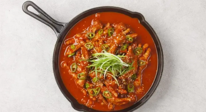 Kurczak w sosie gochujang, posypany sezamem i zieloną cebulką, podany w żeliwnej patelni.