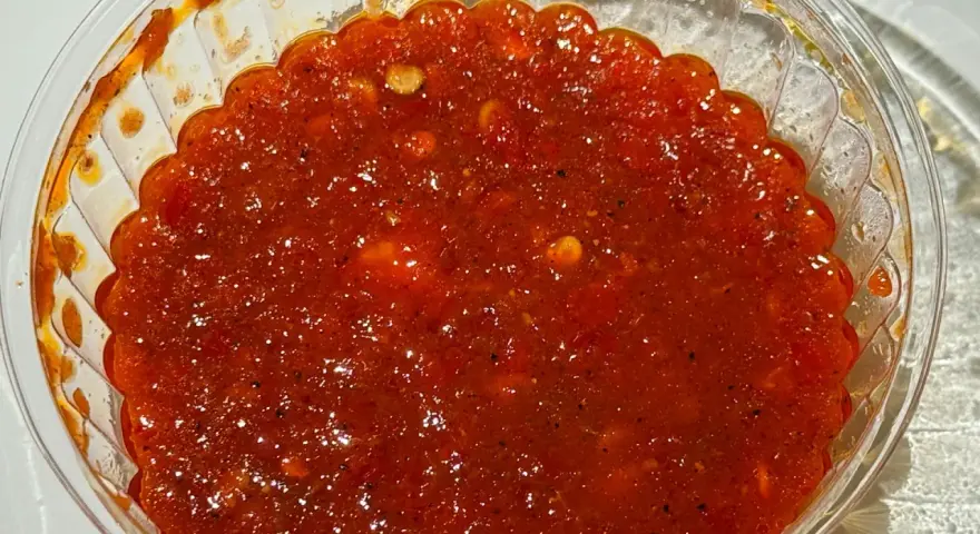 Gęsty, czerwony sos sambal w przezroczystym pojemniku, z widocznymi kawałkami papryczek chili i przypraw.