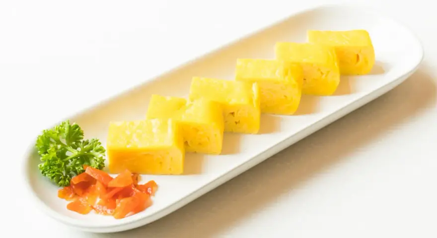 Sześć kawałków puszystego omletu tamago ułożonych na białej tacy, ozdobionej natką pietruszki i pomarańczowym dodatkiem.
