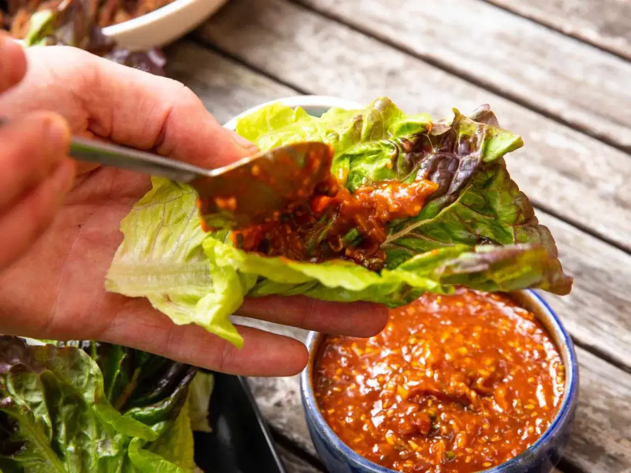 Ręka nakłada łyżką pikantny sos gochujang na liść sałaty. Obok miseczka z sosem.