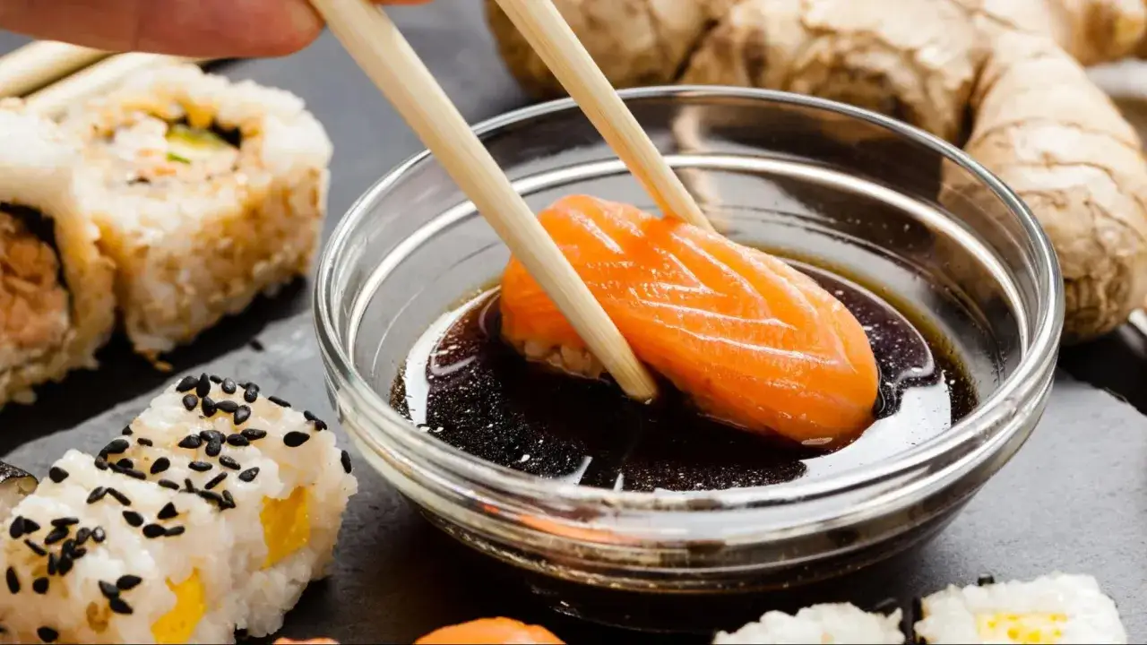 Kawałek łososia zanurza się w sosie do sushi. Obok leżą rolki sushi i imbir.