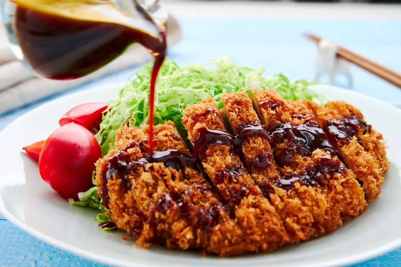 Chrupiący kotlet schabowy polany ciemnym, aromatycznym sosem tonkatsu, podany z sałatką z poszatkowanej kapusty i pomidorkami.