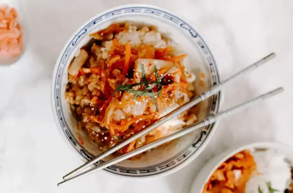 Miseczka ryżu z pikantnym kimchi i startą marchewką, ozdobiona listkami mięty. Obok pałeczki i mała miseczka z dodatkami, jak do koreańskiego banchan.