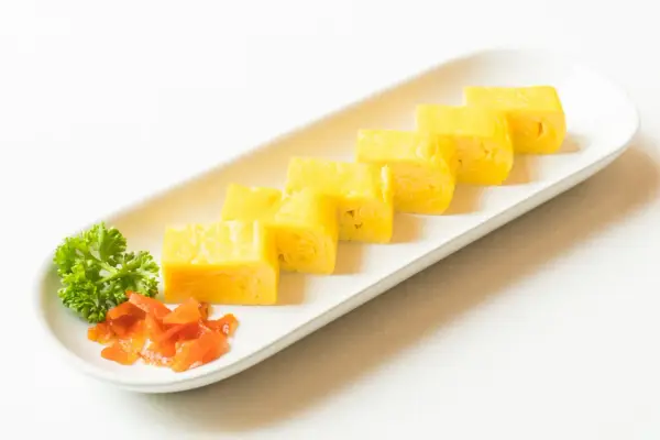 Sześć kawałków puszystego omletu tamago ułożonych na białej tacy, ozdobionej natką pietruszki i pomarańczowym dodatkiem.